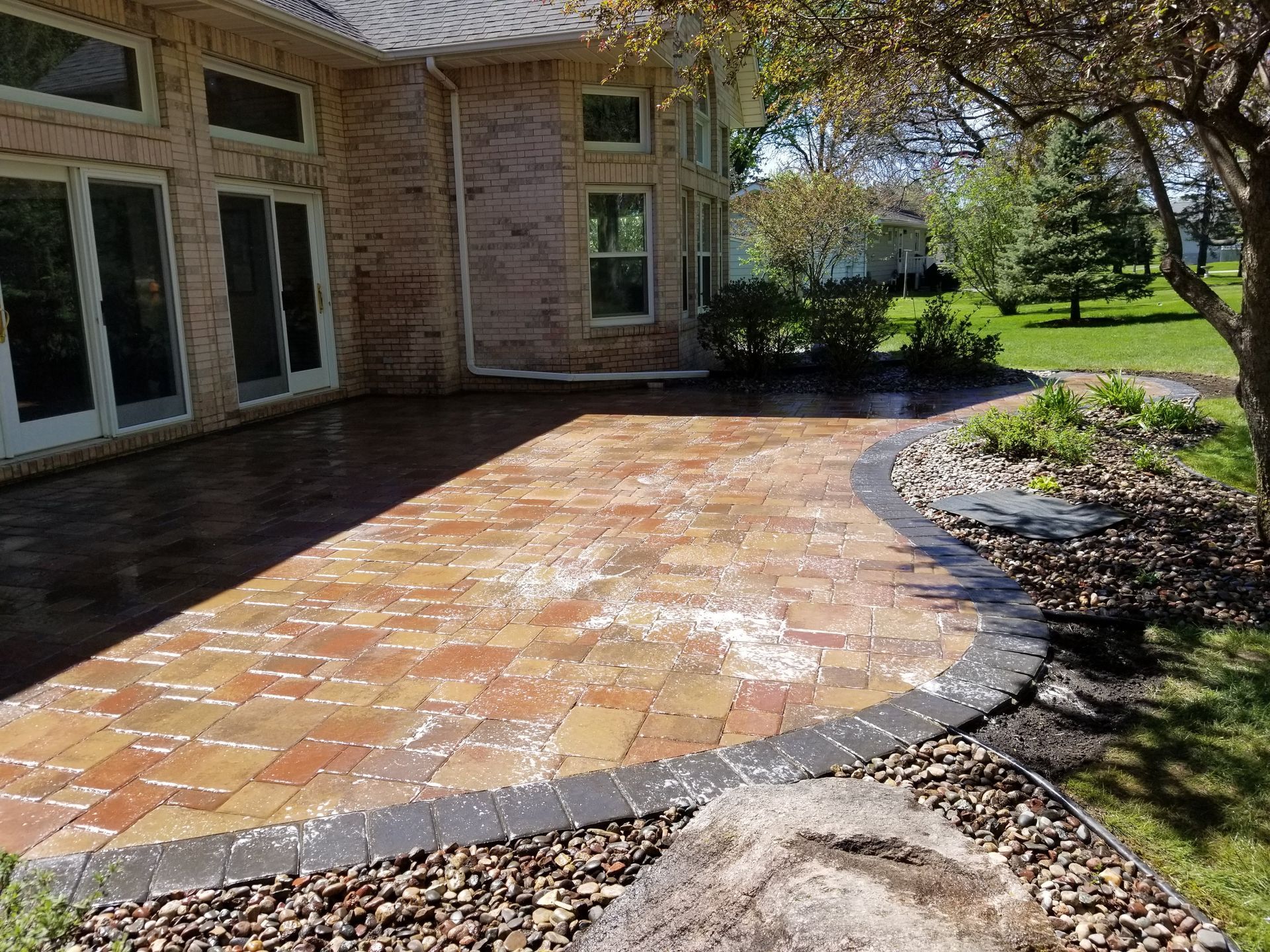 A brick patio with a dark stone border extends from a house into a landscaped yard with trees and mulch.