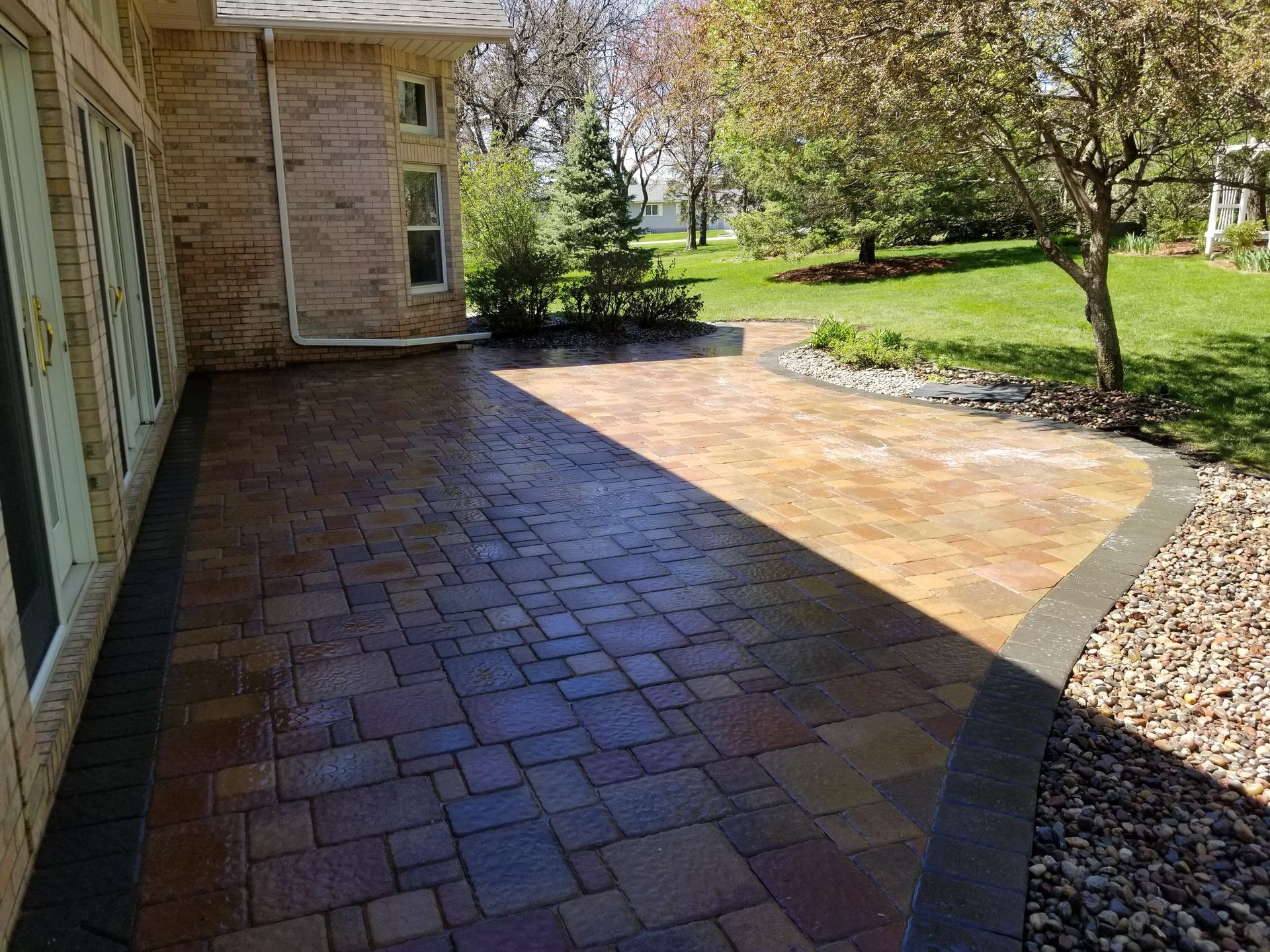A multi-colored paver patio with a dark stone border, extending from a brick house into a green backyard lawn.