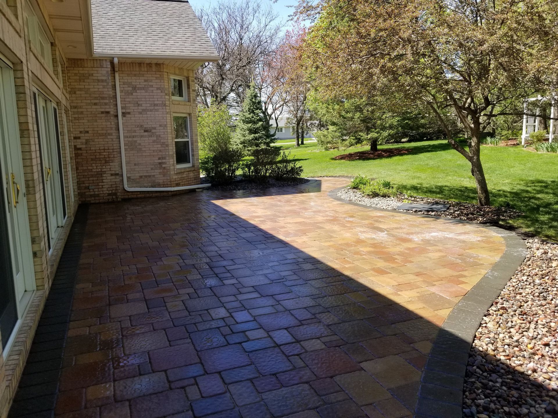 A multi-colored paver patio with a dark stone border extends from a brick house into a sunlit backyard with grass and trees.