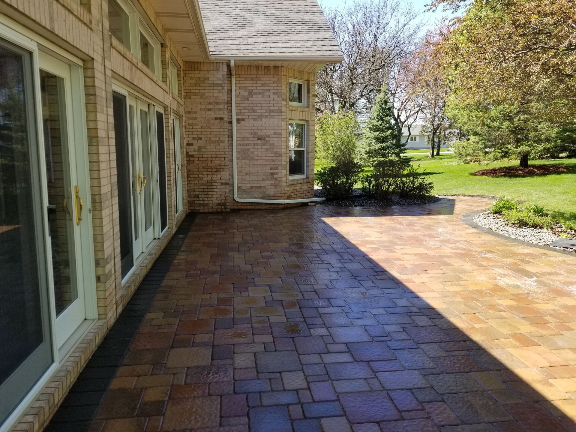 A multi-colored stone patio next to a tan brick house on a sunny day with green grass and trees in the background.