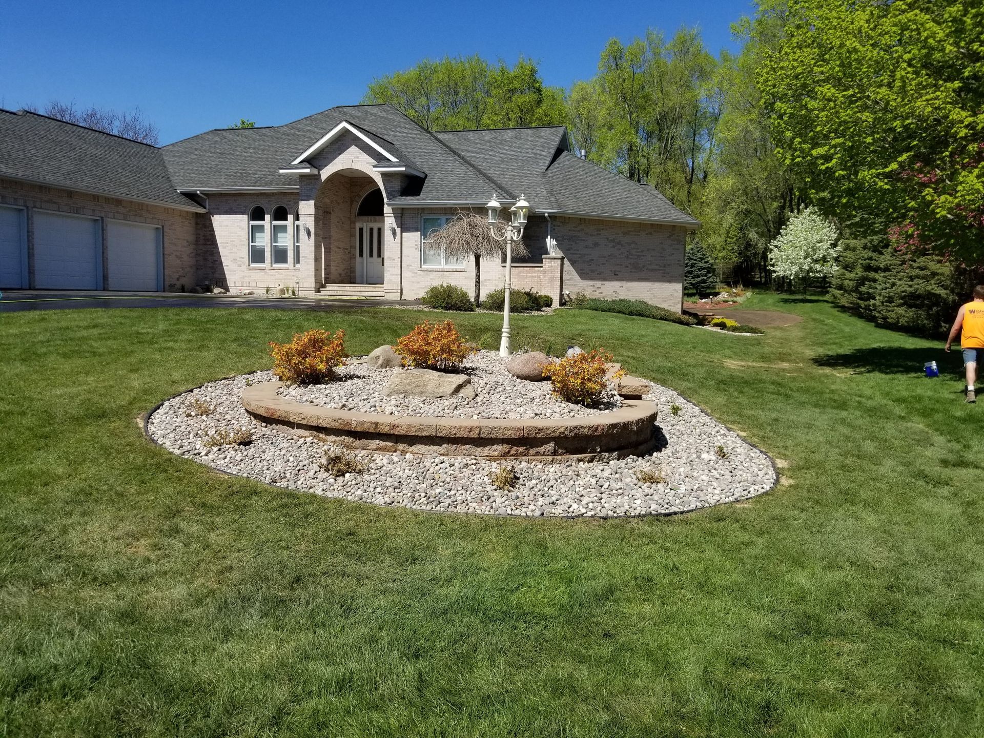 A landscaped stone island in a grassy yard featuring shrubs, a lamp post, and a brick house in the background.