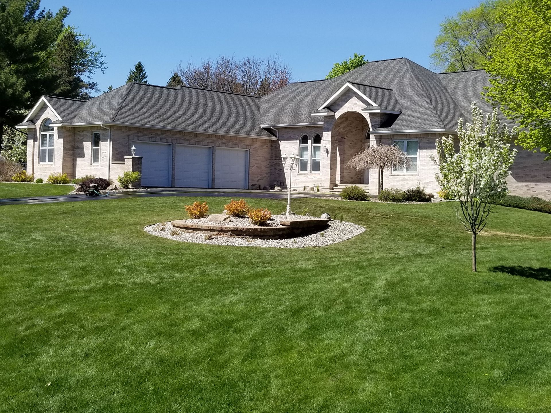 A single-story light brick house with a three-car garage, gray roof, and a landscaped rock bed in a green front yard.