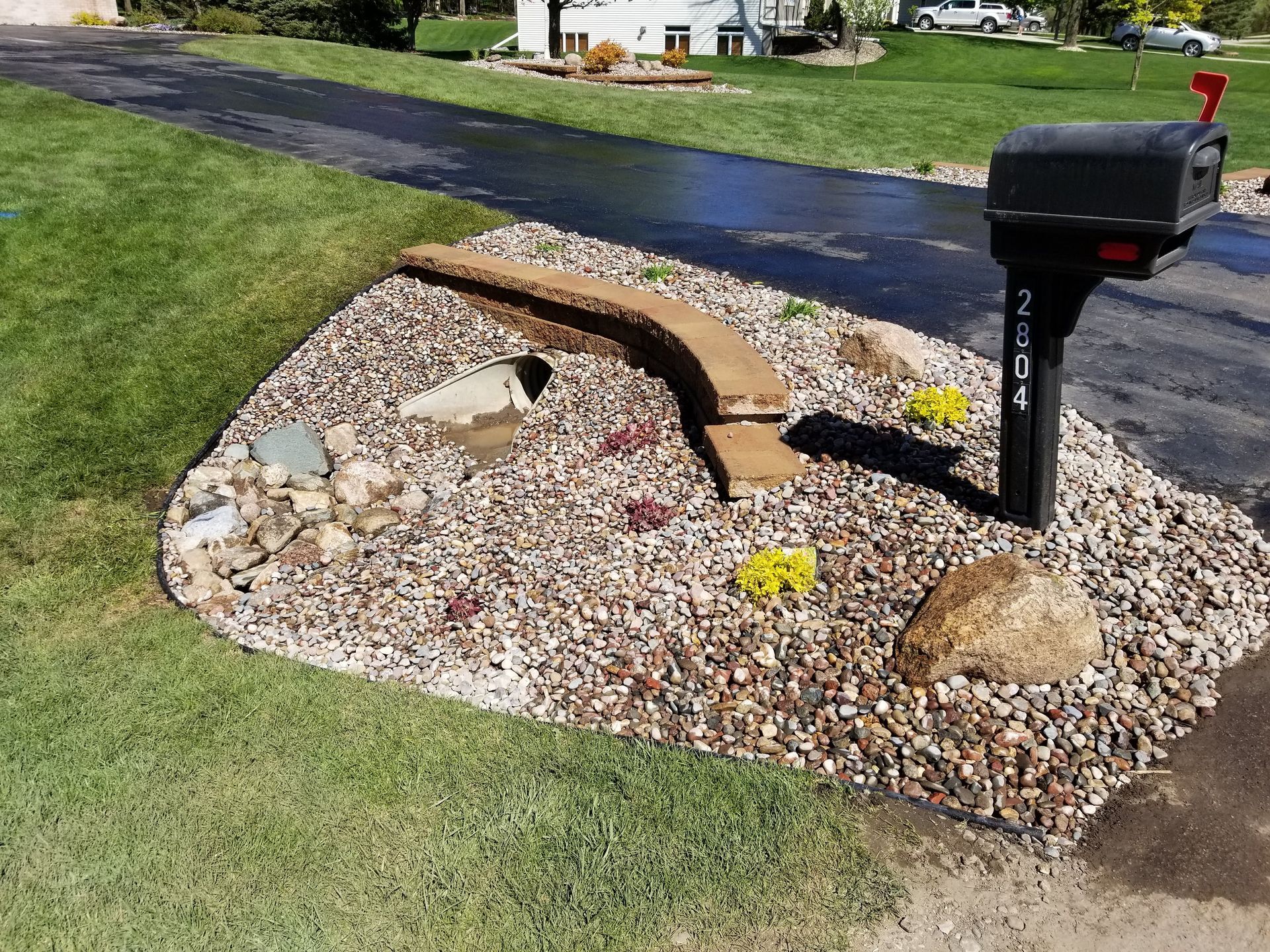 A residential mailbox sits next to a landscaped stone bed with a small retaining wall on a suburban lawn by a driveway.