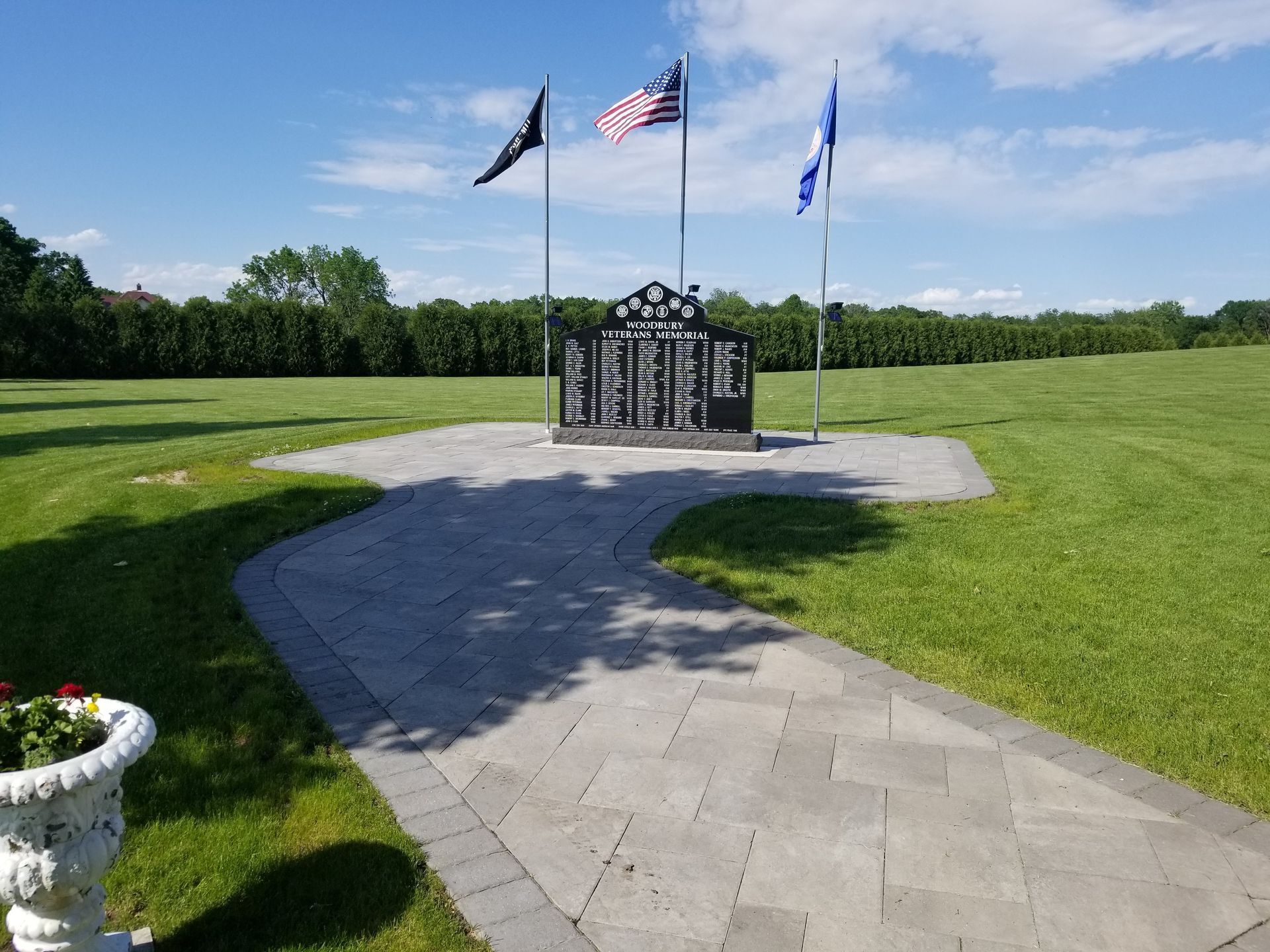 A stone memorial with three flags sits in a green field at the end of a paved walkway, with a flower pot in the foreground.