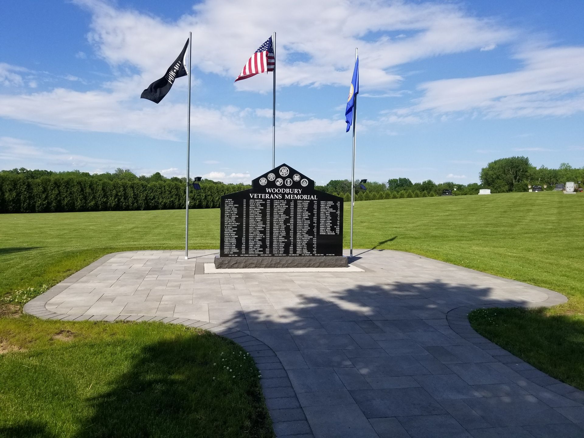 A black granite memorial with names stands in a grassy park, flanked by three flags under a bright blue sky.
