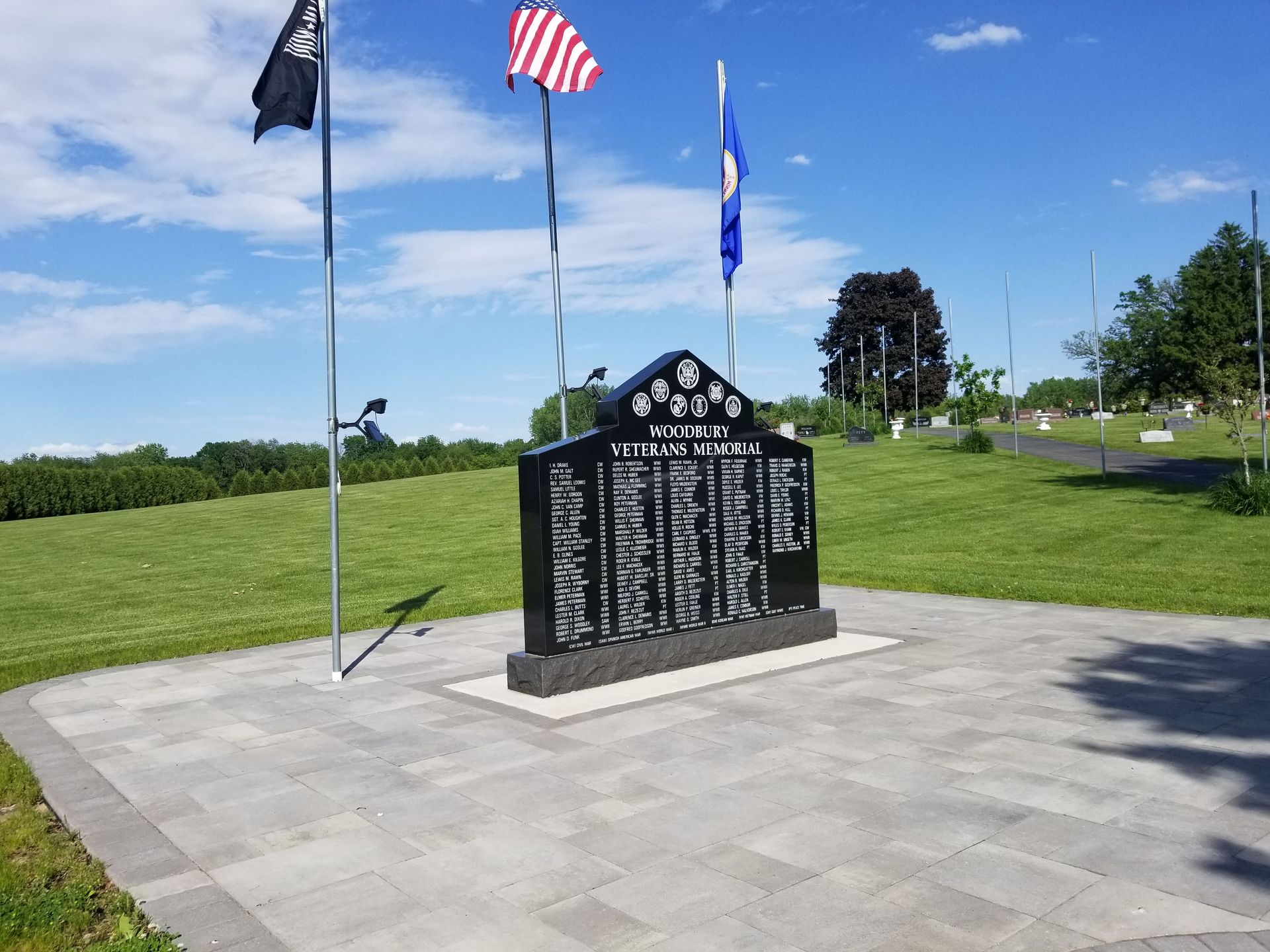 A black stone memorial with engraved names stands on a paved plaza, flanked by three flags under a bright blue sky.