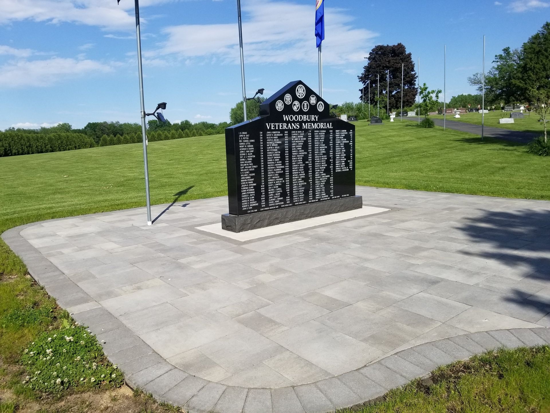 A black granite veterans memorial monument stands on a paved stone plaza in a green, grassy field under a sunny sky.