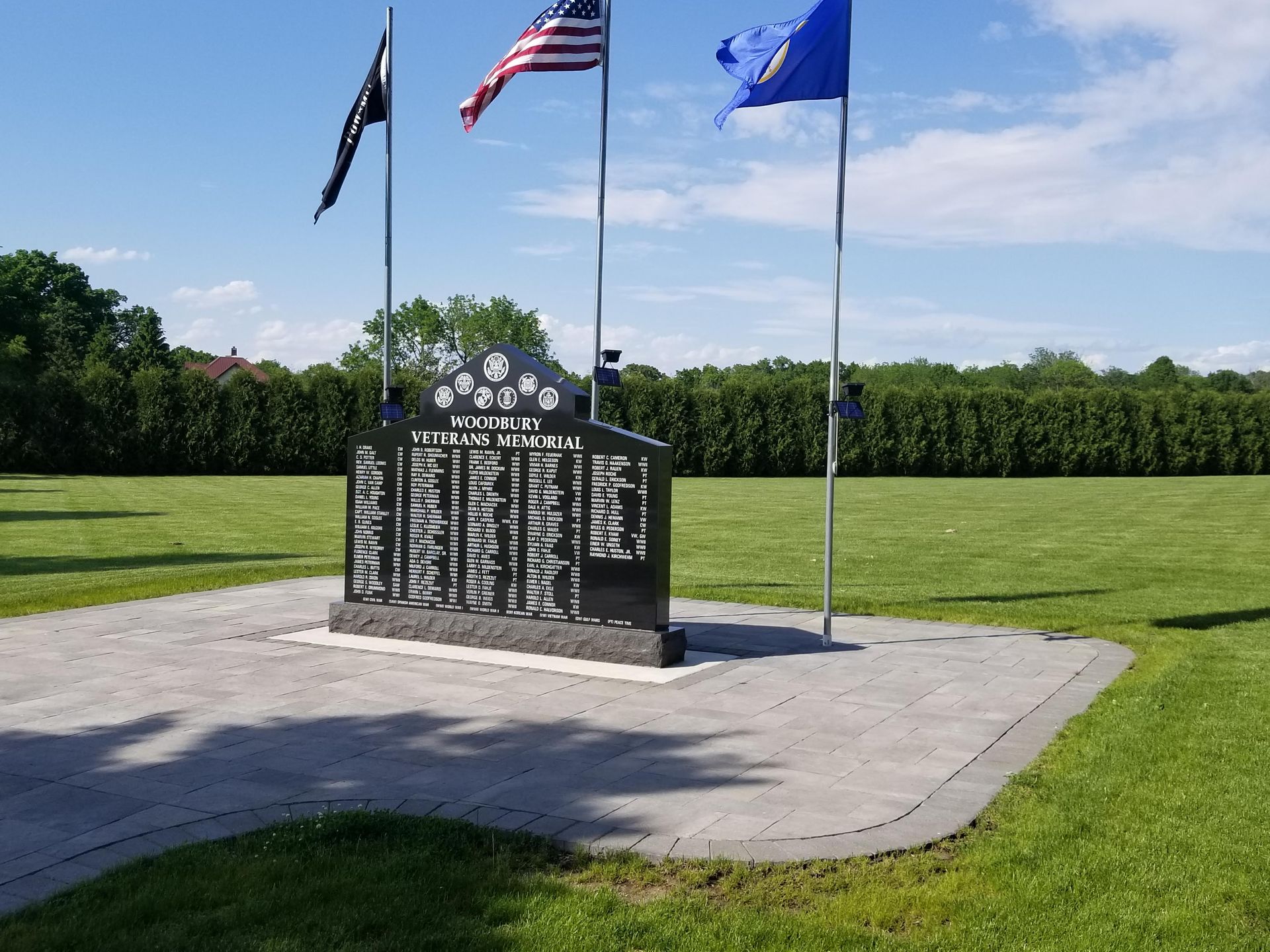 A black granite war memorial monument stands on a paved stone plaza, flanked by three flags in a grassy park setting.