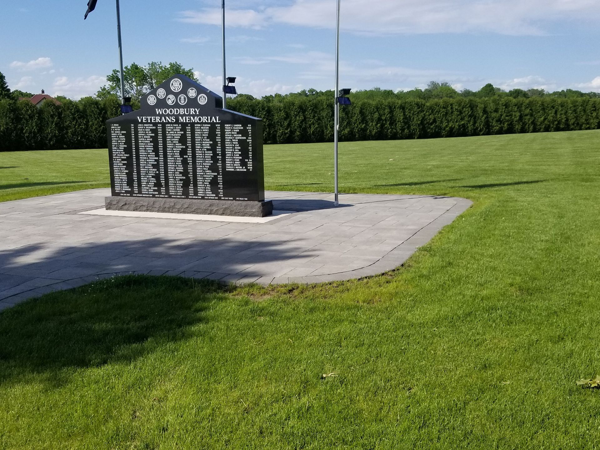 A black stone veterans memorial with engraved names stands on a paved base in a grassy field under a blue, sunny sky.