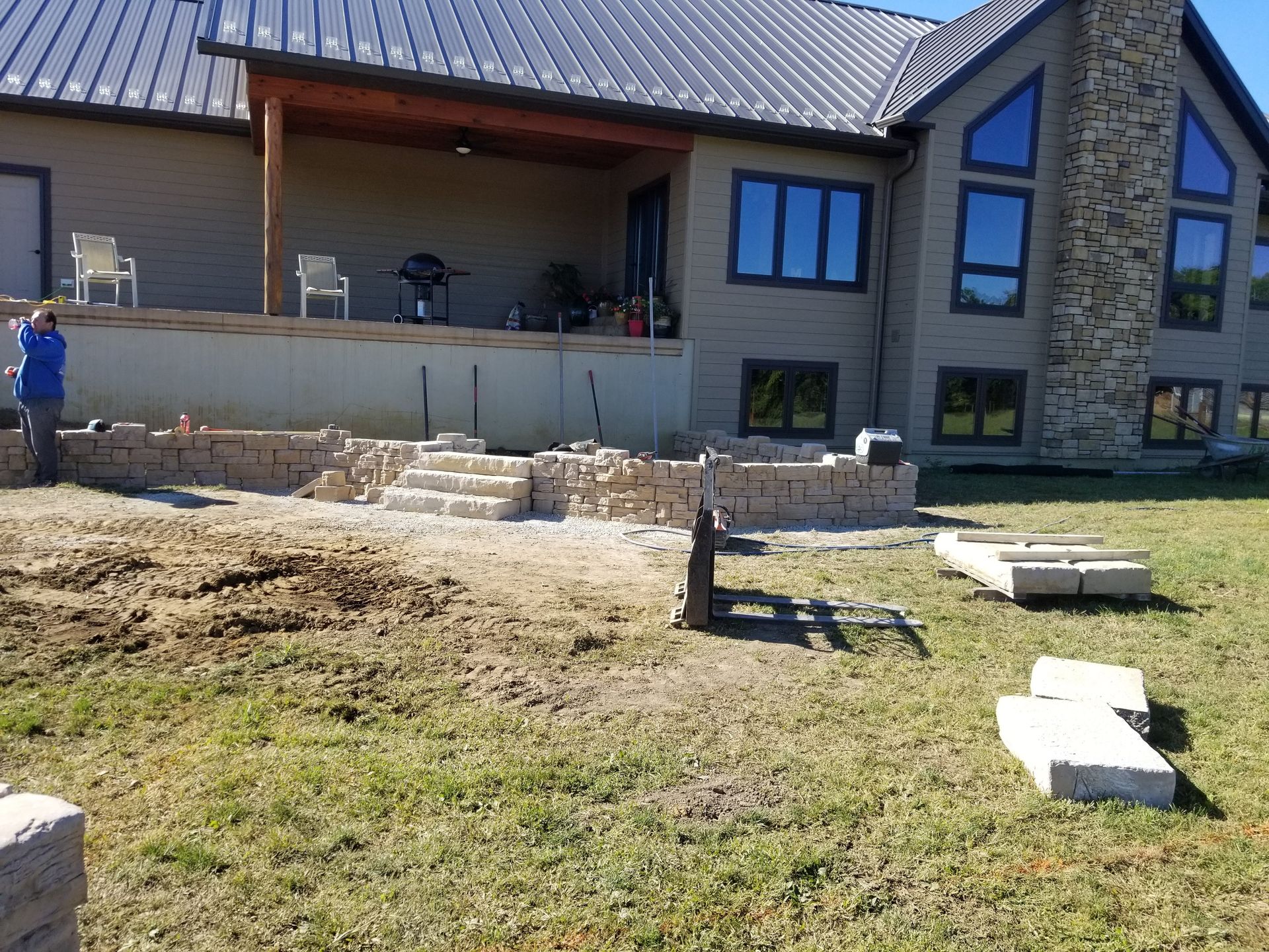 A person stands near a stone retaining wall under construction in the backyard of a modern home with a metal roof.