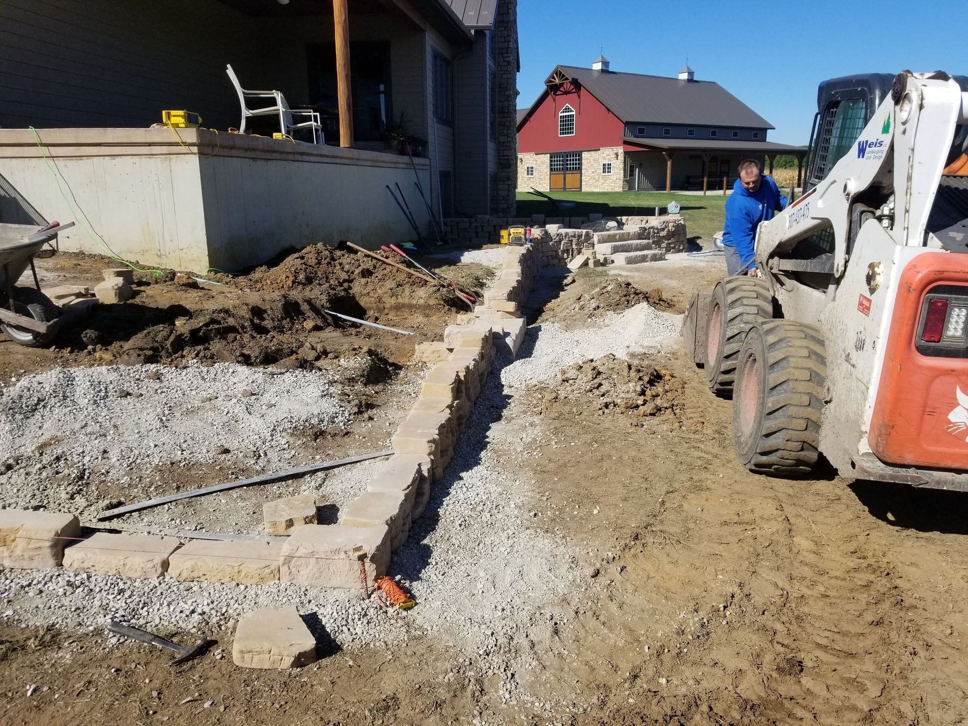 A person operates a skid-steer loader near a stone retaining wall under construction in a yard with a red barn nearby.