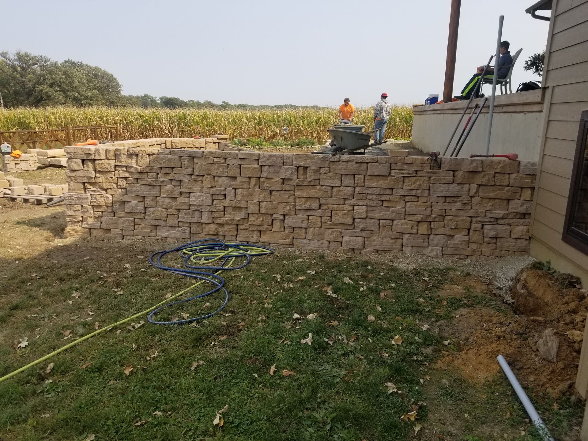 Two workers build a stone retaining wall in a yard next to a field, while another person sits on a nearby porch.