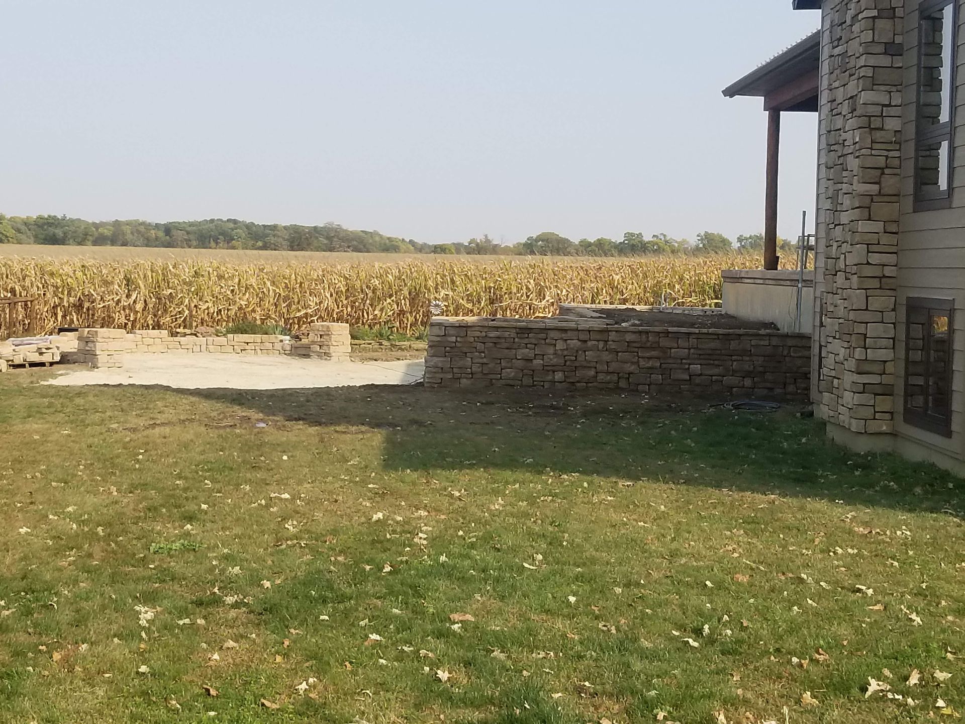 A stone patio and retaining wall extend from the side of a house toward a vast, dry cornfield under a hazy sky.