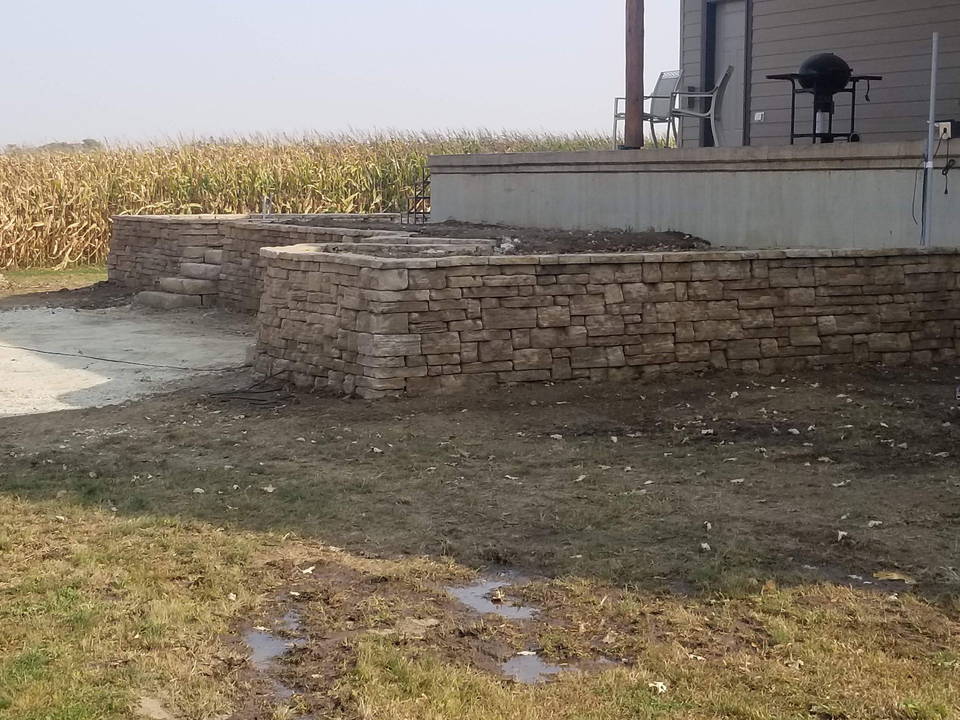 A beige stone retaining wall sits in front of a house porch next to a cornfield under a hazy sky.