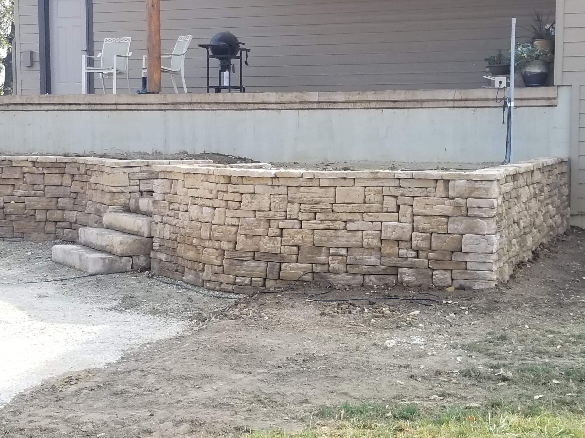 A newly constructed natural stone retaining wall with integrated steps in front of a house patio with chairs and a grill.