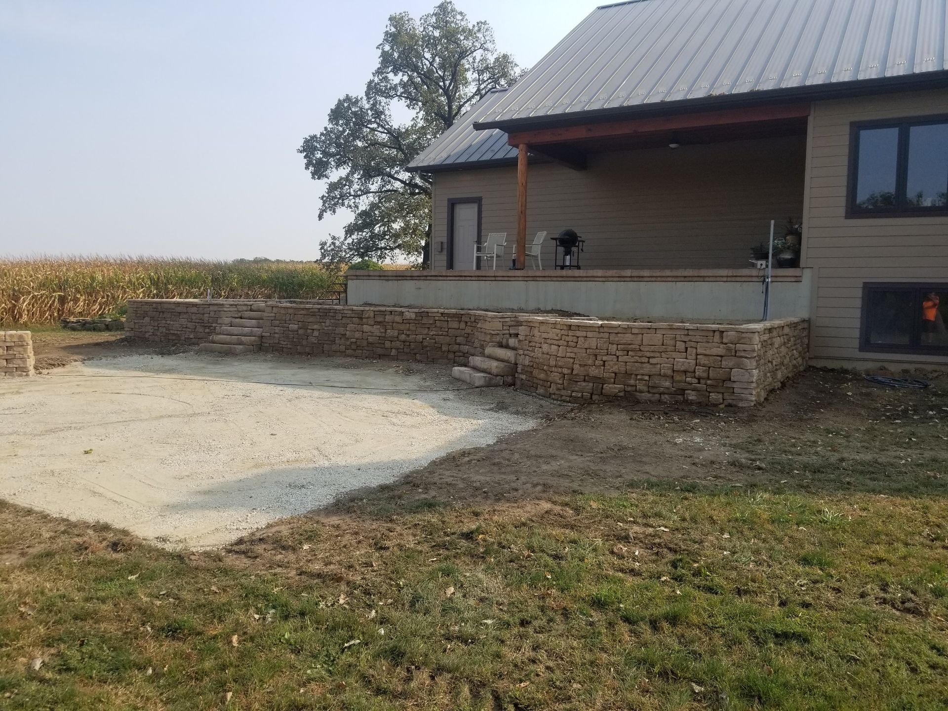A stone retaining wall with steps leads up to a covered patio on a house next to a field of crops.