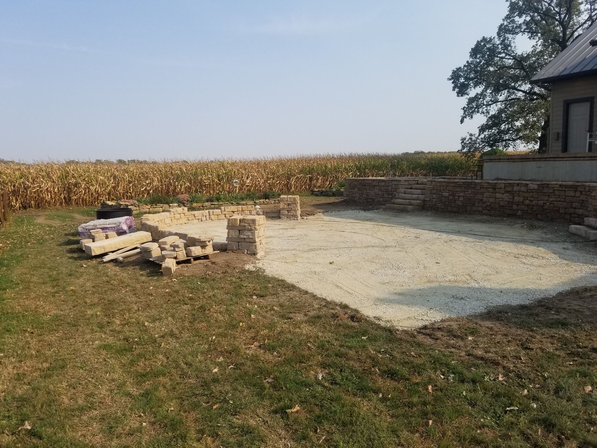 A gravel patio under construction with stone pillar bases beside a cornfield and a house.