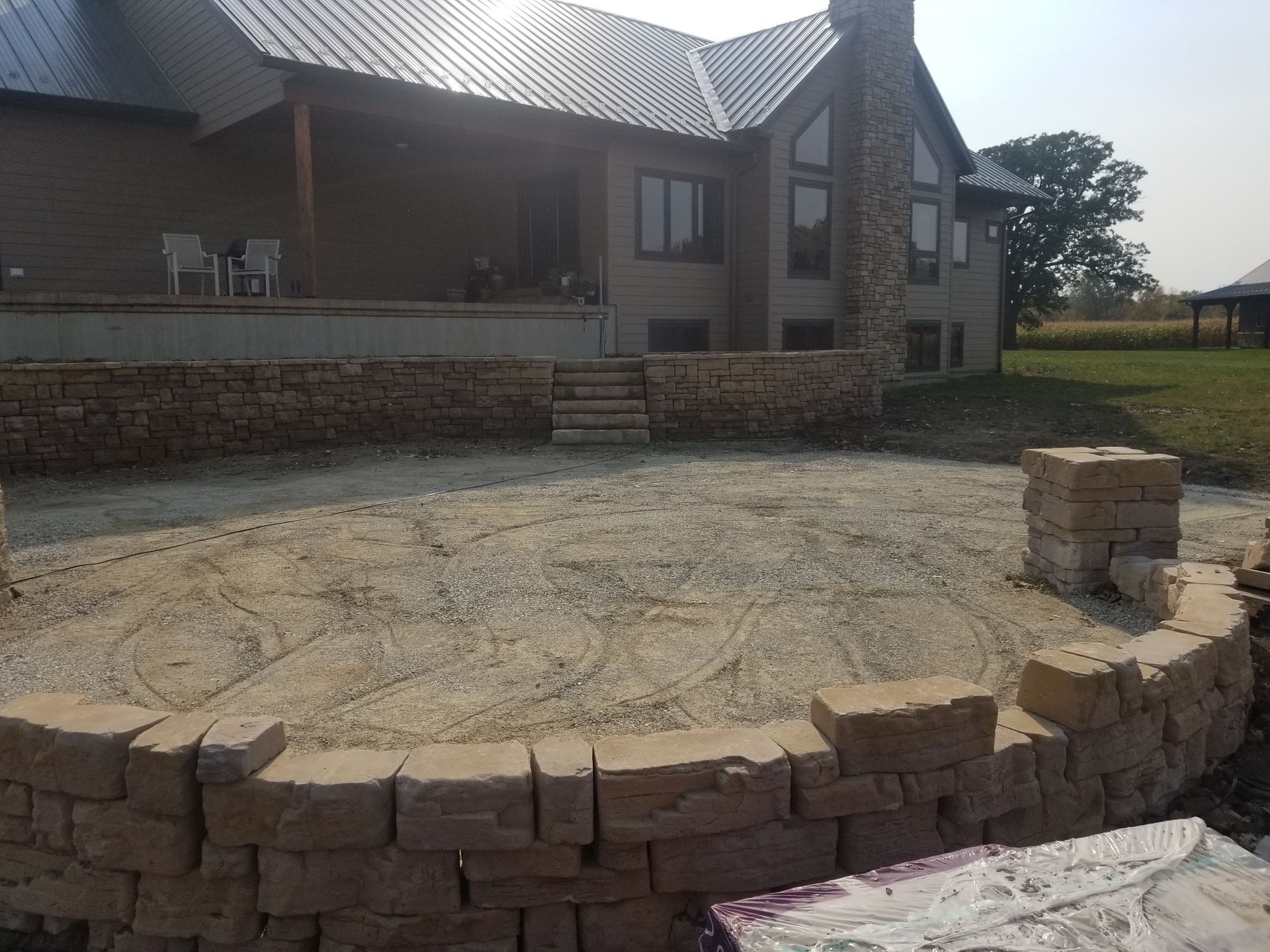 A tan stone retaining wall curves around a gravel patio area in front of a house with a stone chimney.