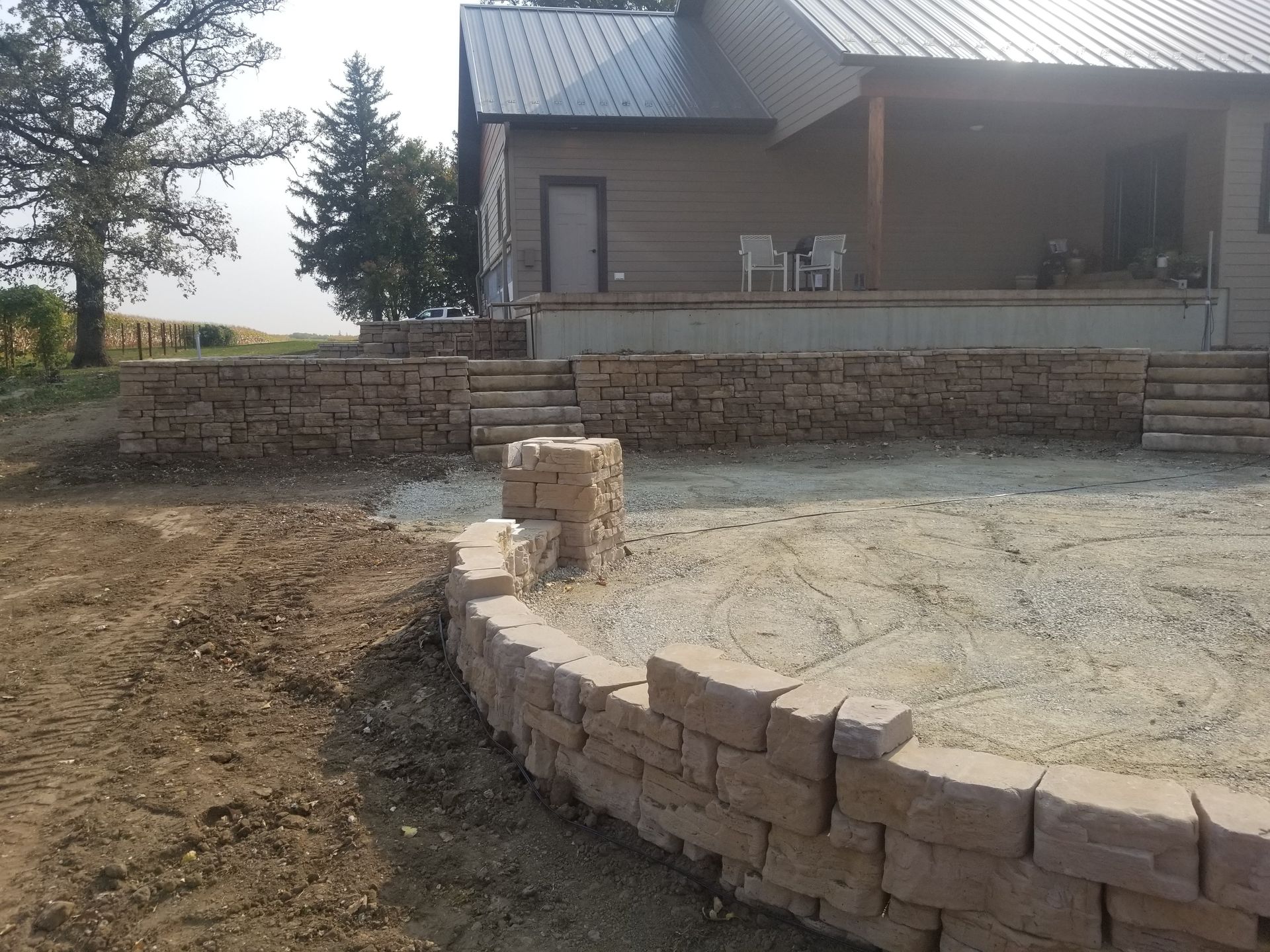 A stone retaining wall curves around a gravel patio in front of a house with a porch.