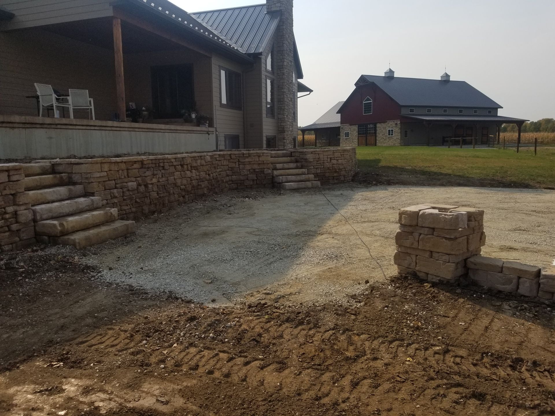 A stone retaining wall and staircase under construction in front of a house, with a red barn in the background.
