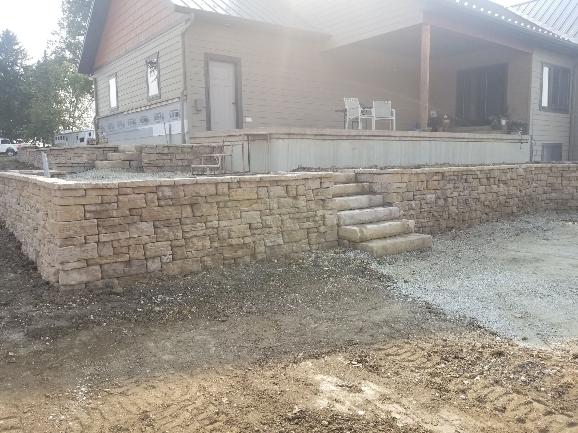 A stone retaining wall with steps leads up to a tan house with a porch under construction.
