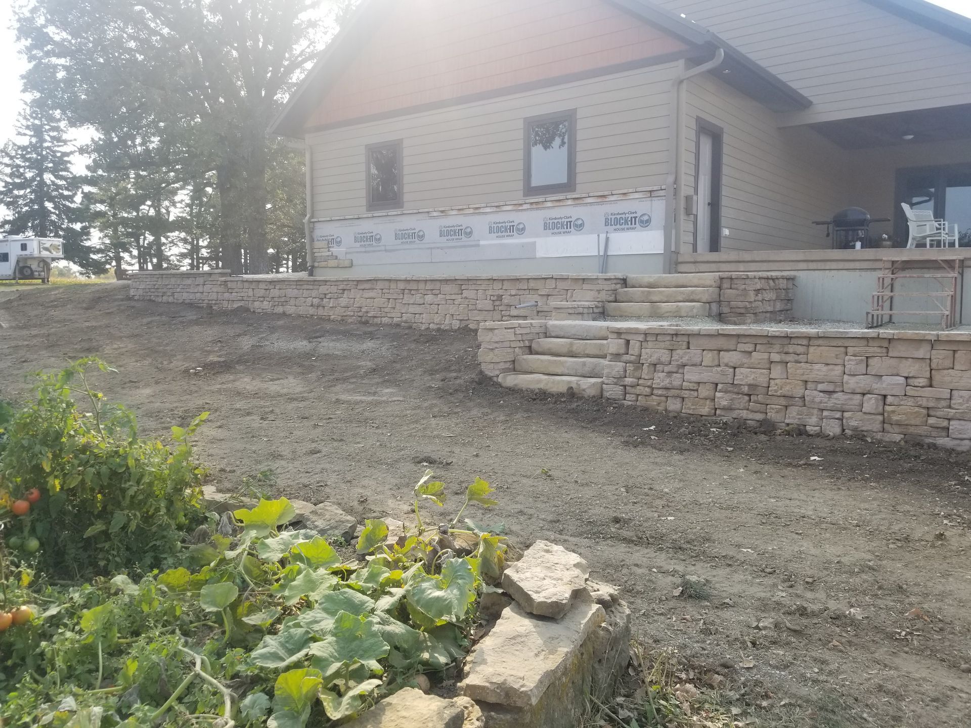 A beige house with unfinished exterior walls, featuring a tiered natural stone retaining wall and steps in the yard.