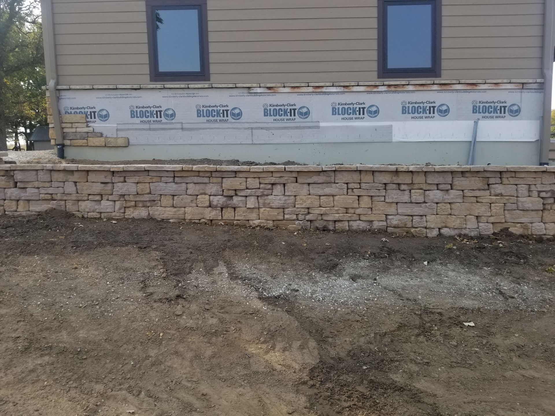 Tan siding house under construction featuring a stone retaining wall base with exposed white vapor barrier on the foundation.