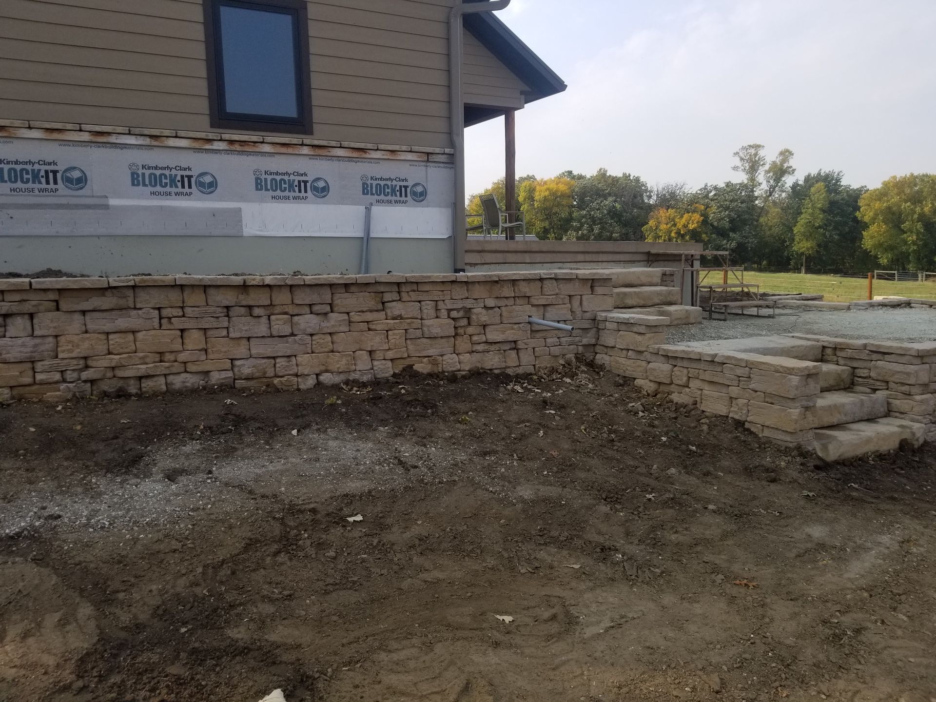 A stone retaining wall under construction alongside a house with tan siding and a gravel yard.