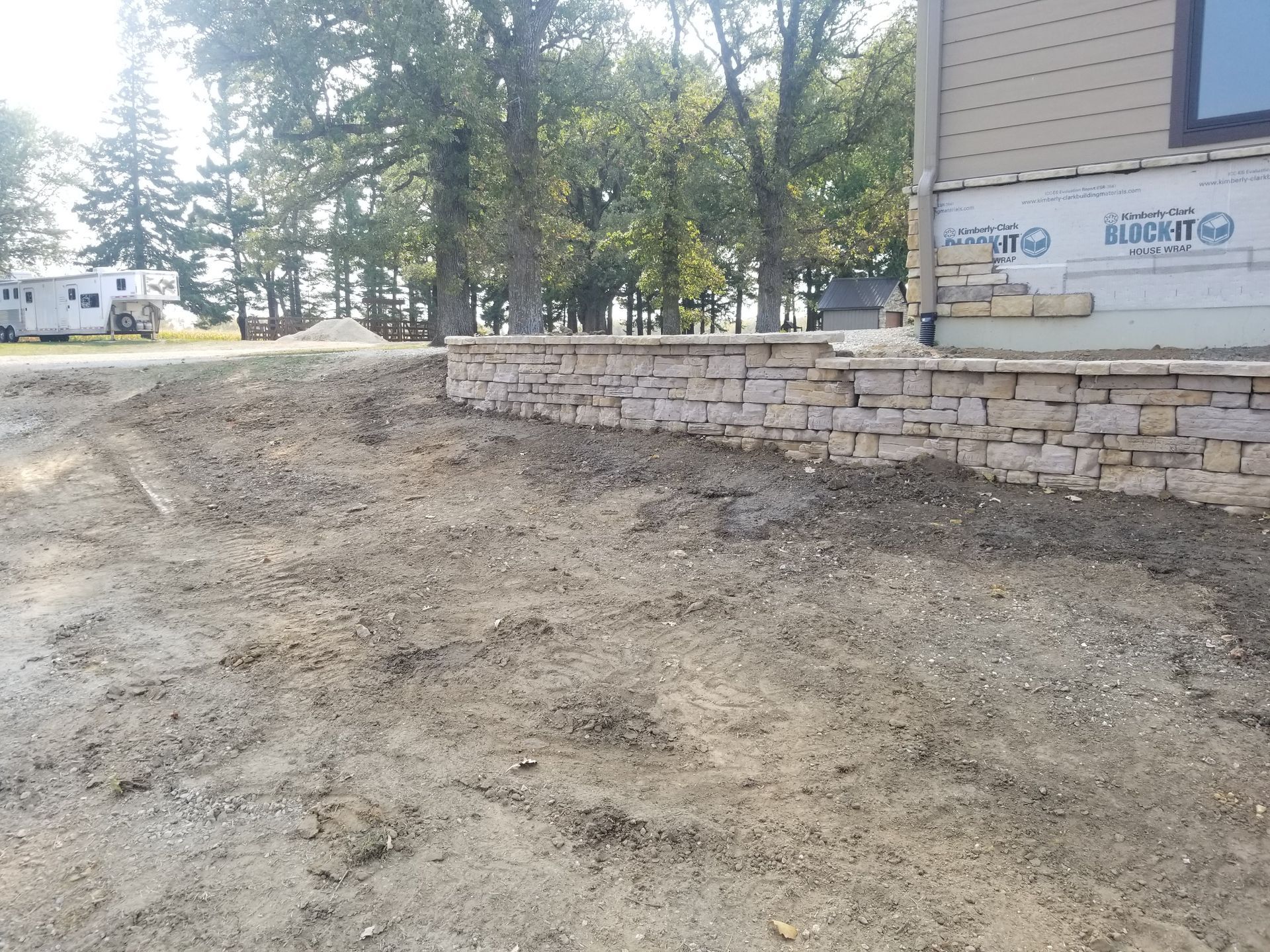 A stone retaining wall stands in front of a house under construction, with dirt ground and trees in the background.