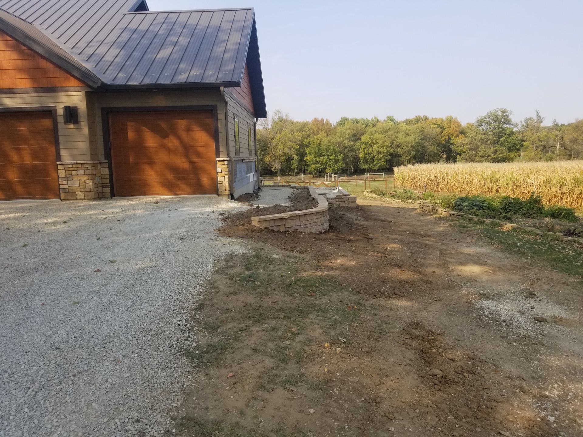 A gravel driveway leads to a house with a stone retaining wall in front of a rural field of dry corn.