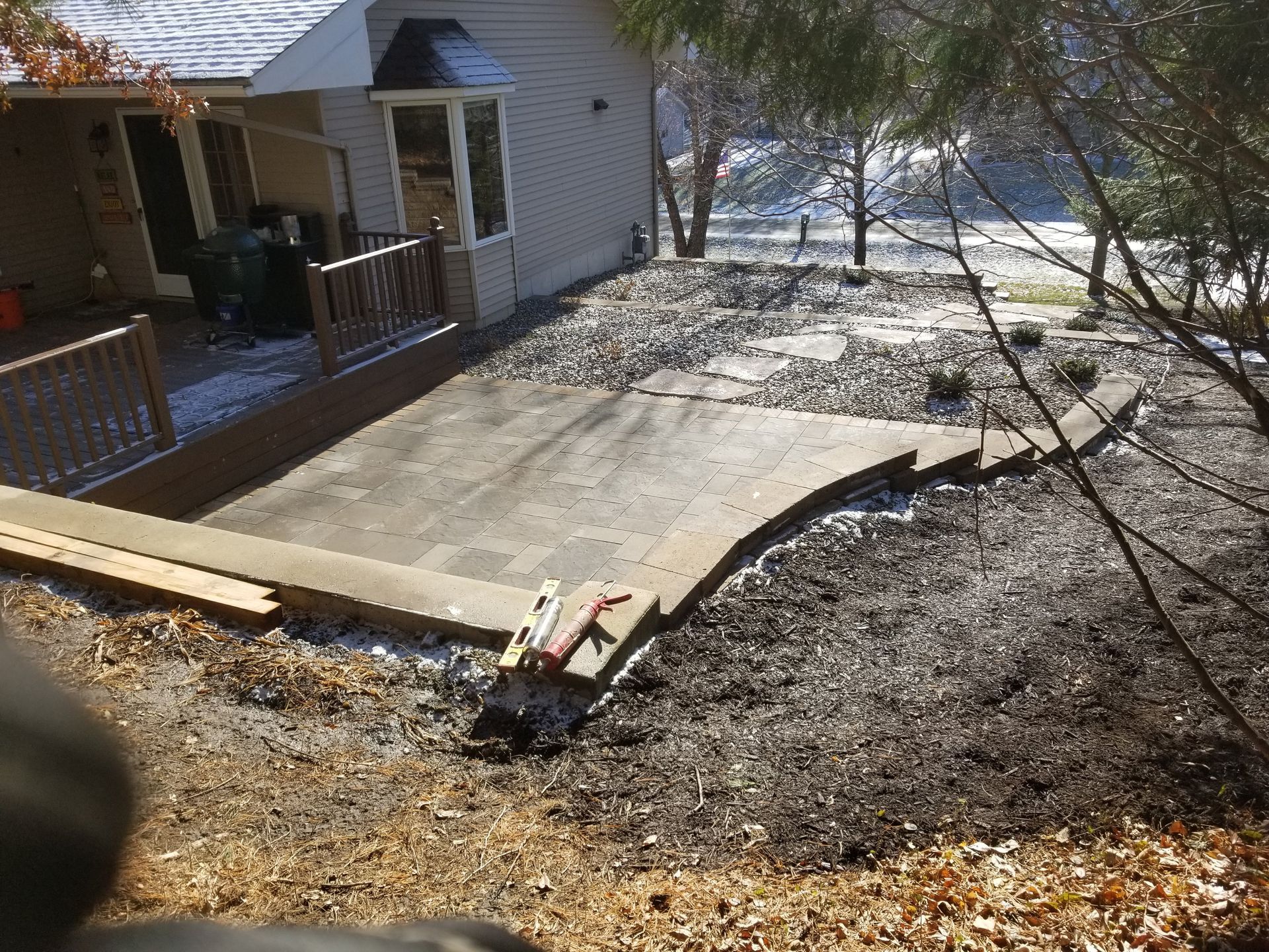 A newly constructed stone paver patio sits next to a house deck, with a small stone retaining wall and surrounding soil.