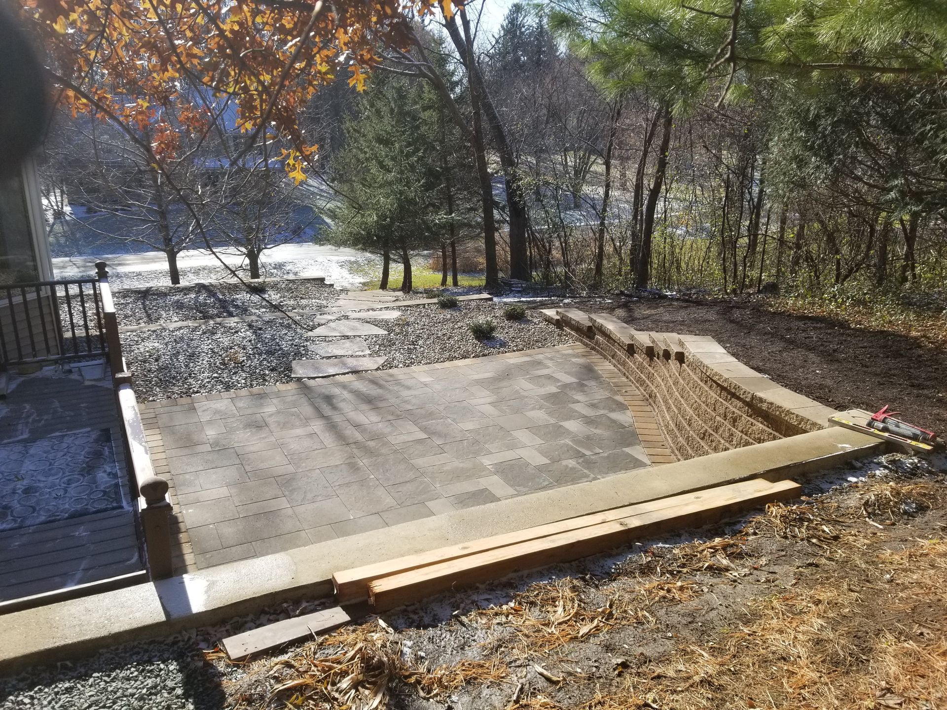 A backyard area with a newly installed gray paver patio, a retaining wall, and a stone path leading toward a wooded yard.