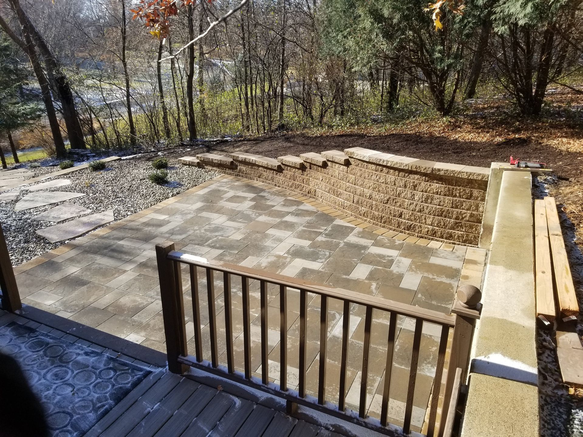 A stone patio under construction, with a wood railing and a retaining wall, set in a wooded backyard.