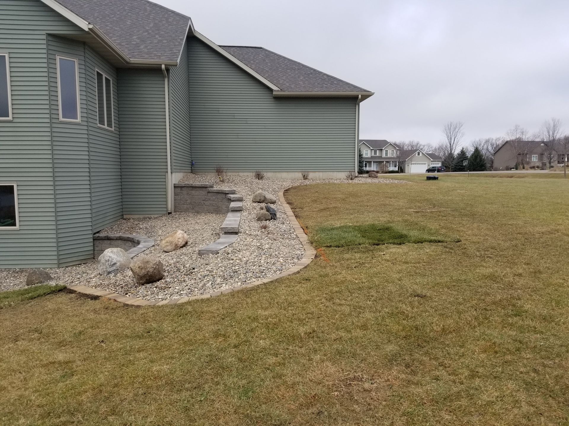A sage green house with a light-colored rock landscaping bed and a stacked stone border along the foundation.