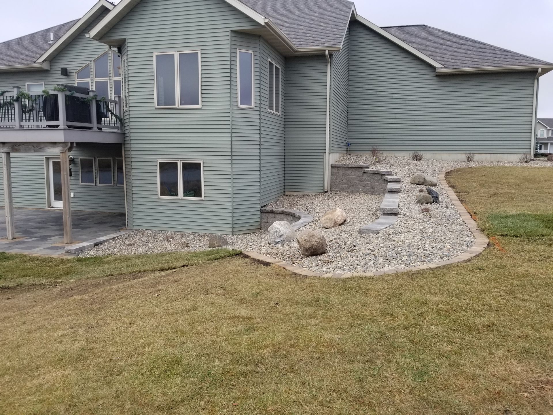 Back of a green-sided house with a raised wooden deck, patio, and a landscaped rock garden bed along the foundation.
