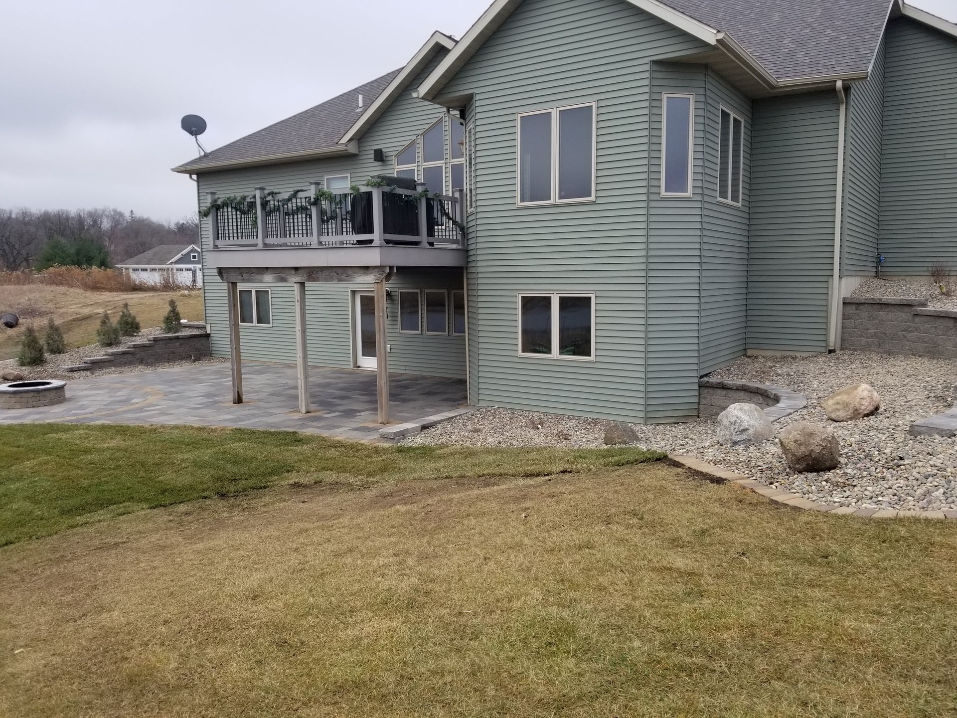 A light green house with a raised wooden deck over a stone patio, surrounded by grass and landscaping rocks.