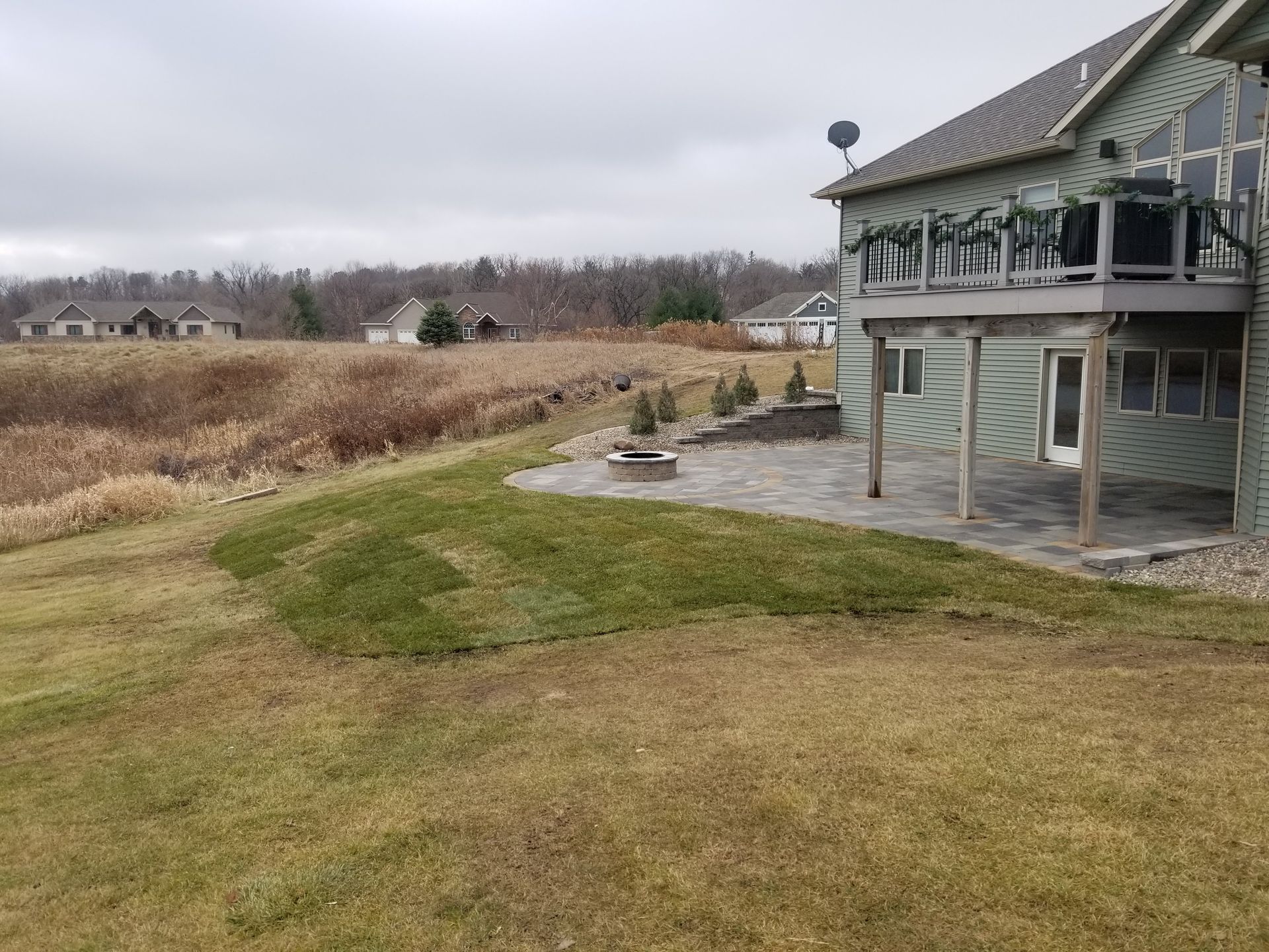 A green house with a raised wooden deck and a stone patio featuring new sod in the backyard against a cloudy sky.