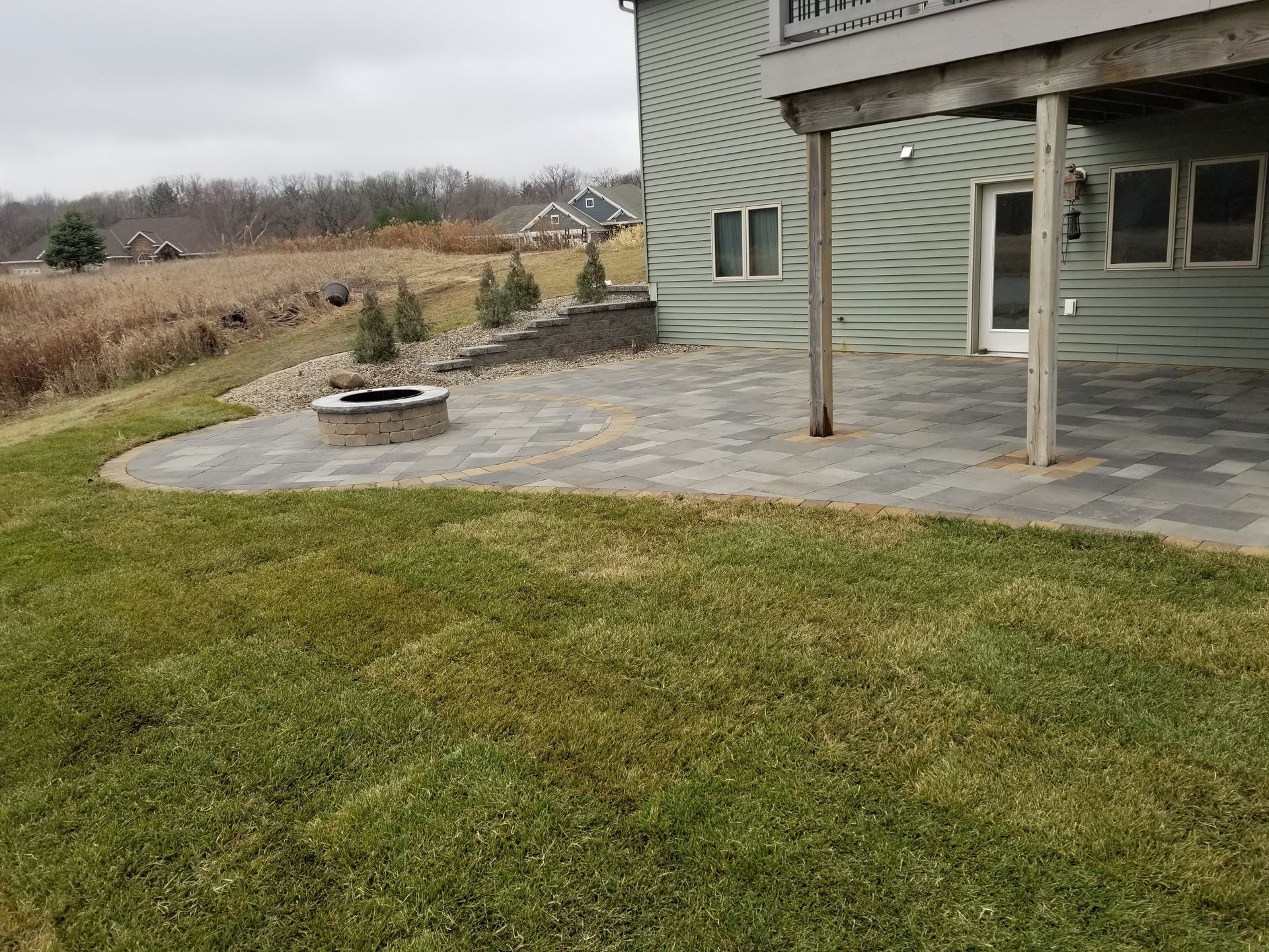 A paved patio with a stone fire pit attached to the back of a green-sided house, overlooking a grassy field.