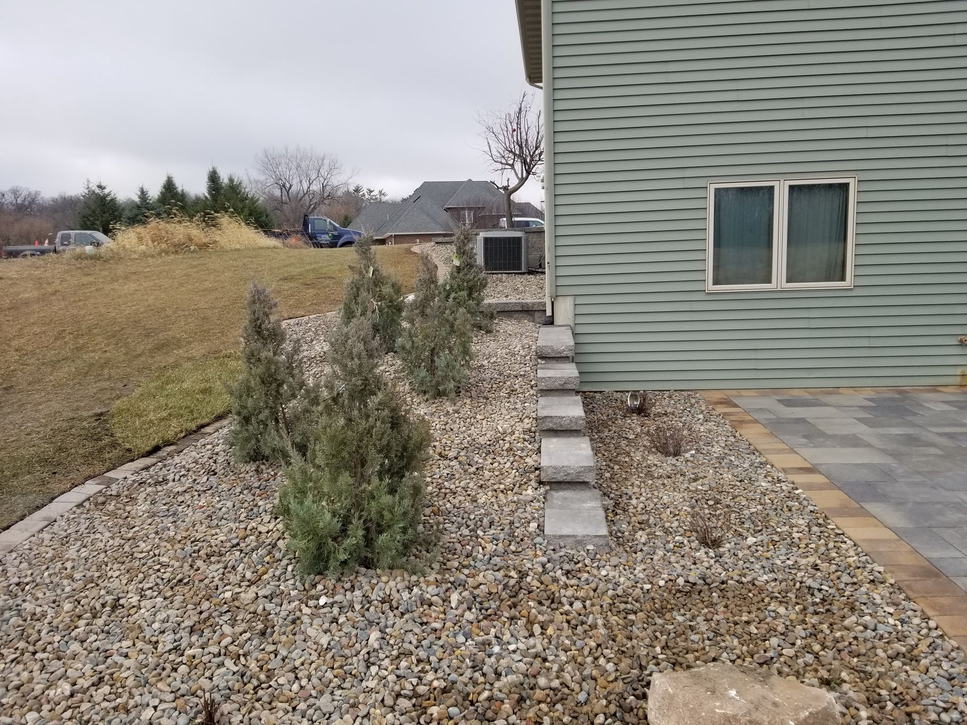 A gravel yard beside a house with green siding features a row of small evergreen trees and a stone block path.