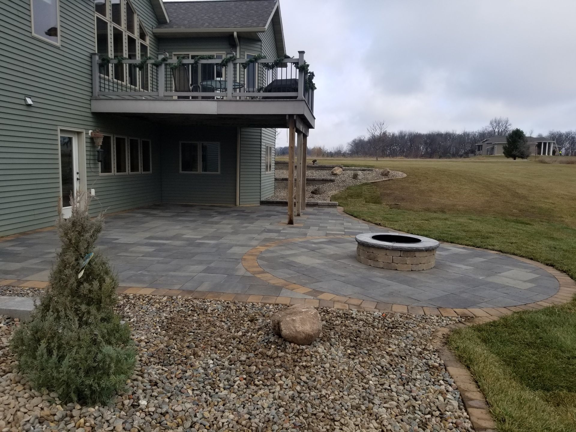 A stone patio with a built-in fire pit sits beside a sage-green house with an elevated deck overlooking a grassy field.