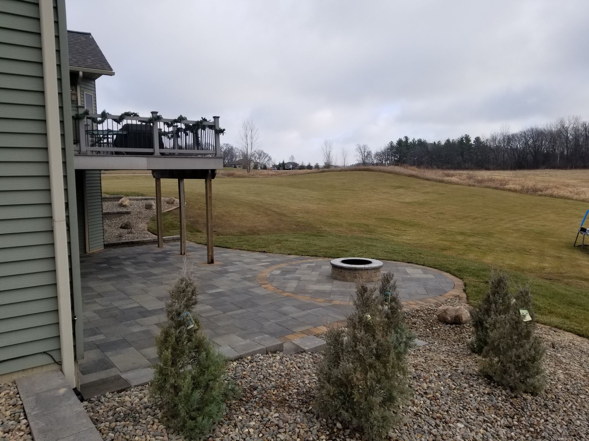 A patio with a stone fire pit under a deck next to a gray house, overlooking an open, grassy field on an overcast day.