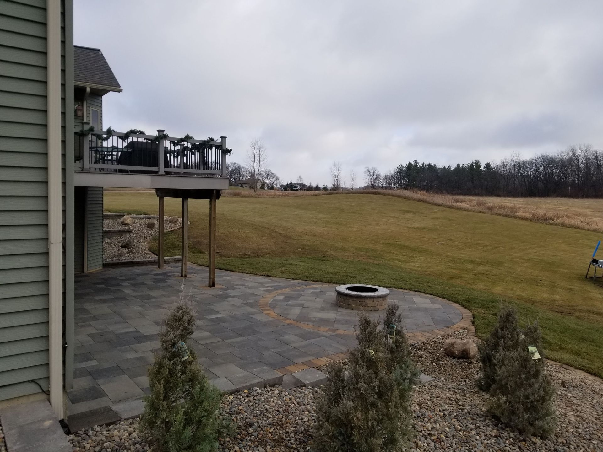 A stone patio with a built-in fire pit sits beneath a wooden deck beside a house, overlooking an expansive, grassy field.