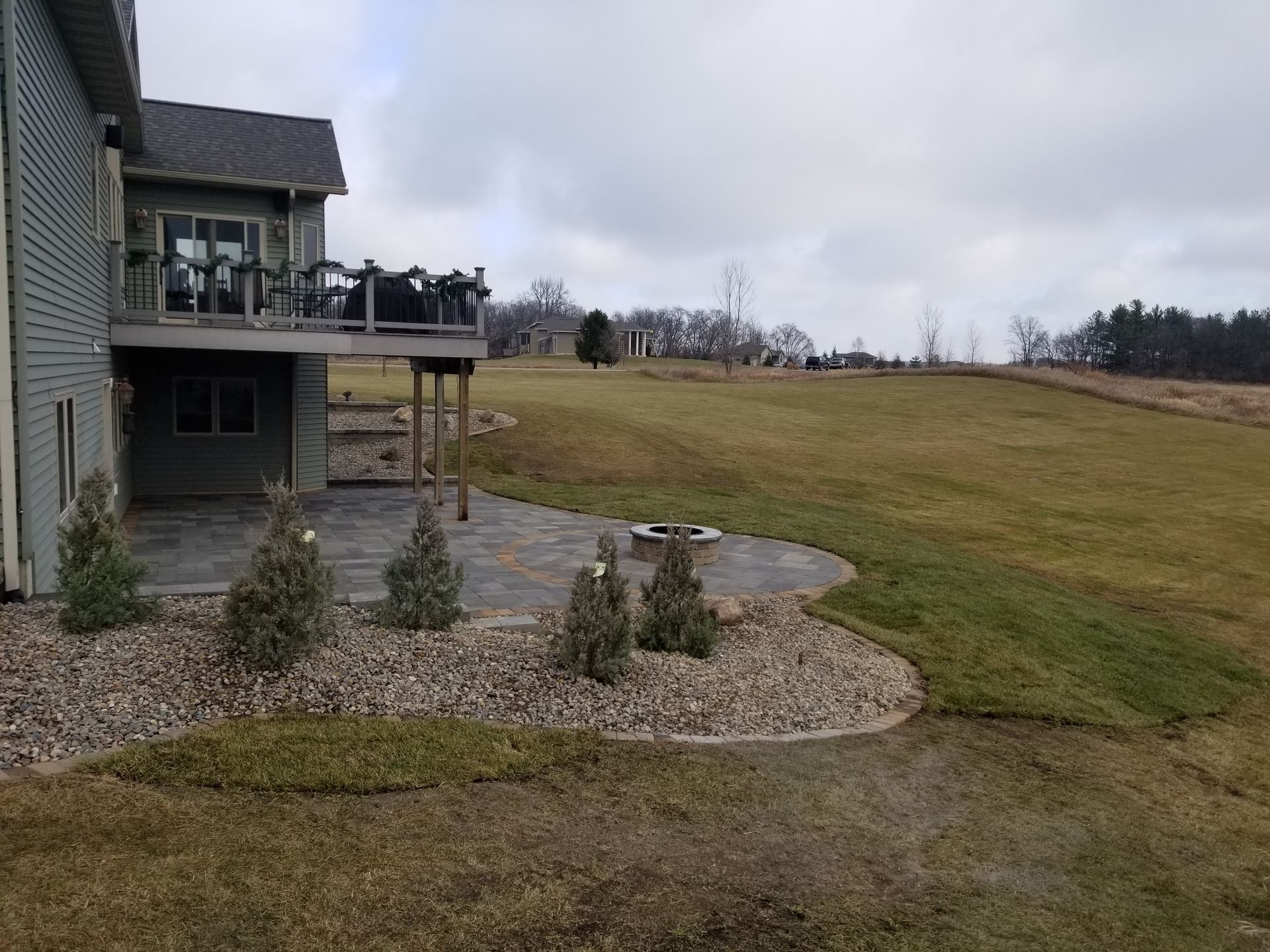 A two-story house with a wooden deck overlooks a paved stone patio, a small landscaped garden, and an open, grassy field.