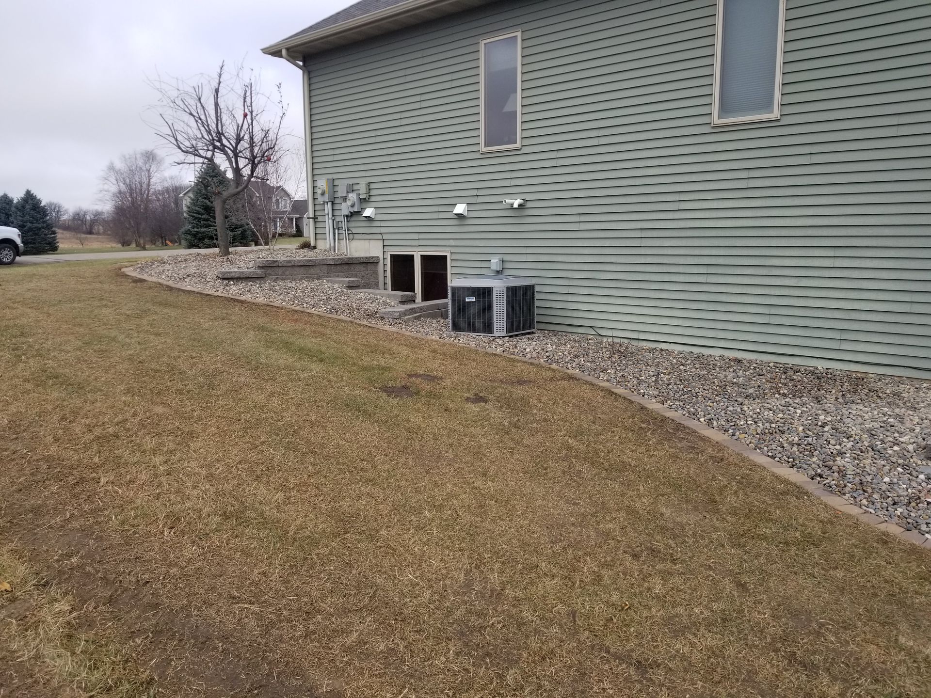 A side view of a house with green siding, featuring a central window, an air conditioning unit, and a gravel yard.