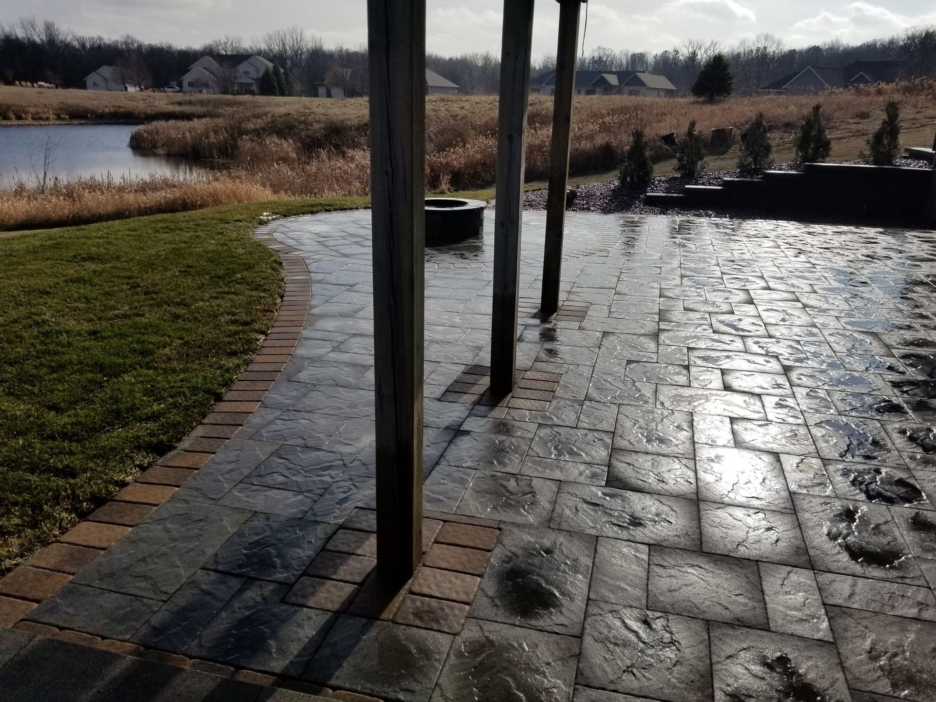 A paved backyard patio with wooden support beams overlooking a pond and grassy field under a bright, sunny sky.