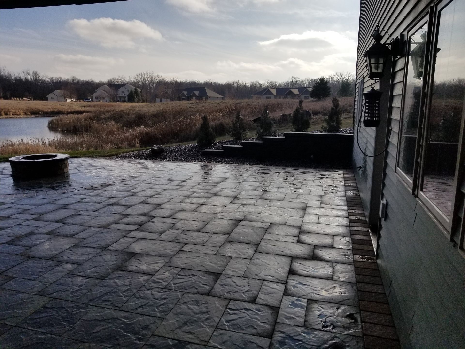 A stone patio with a fire pit overlooking a pond and wetlands on a sunny day, attached to a house with siding.