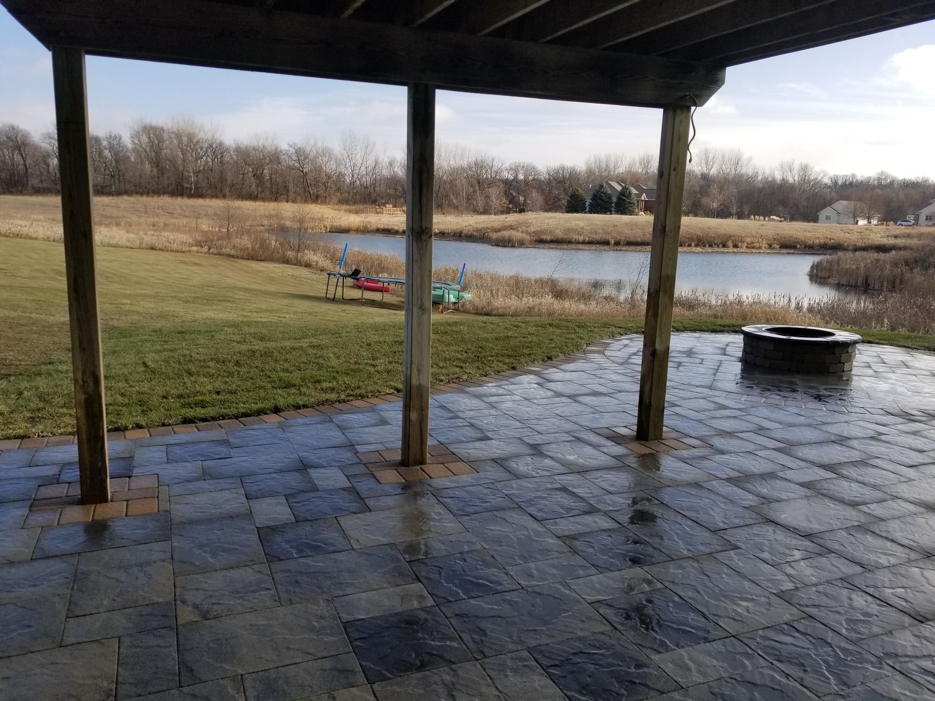 A patio with gray pavers and a fire pit overlooks a pond and grassy field under a sunny sky, viewed from a covered porch.
