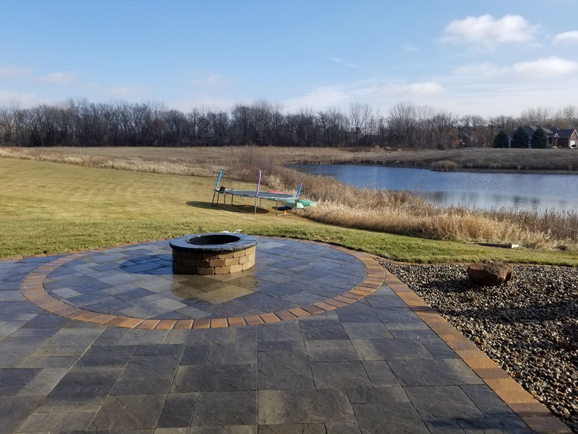 A stone patio with a central circular fire pit overlooking a grassy lawn, a small pond, and a distant treeline.