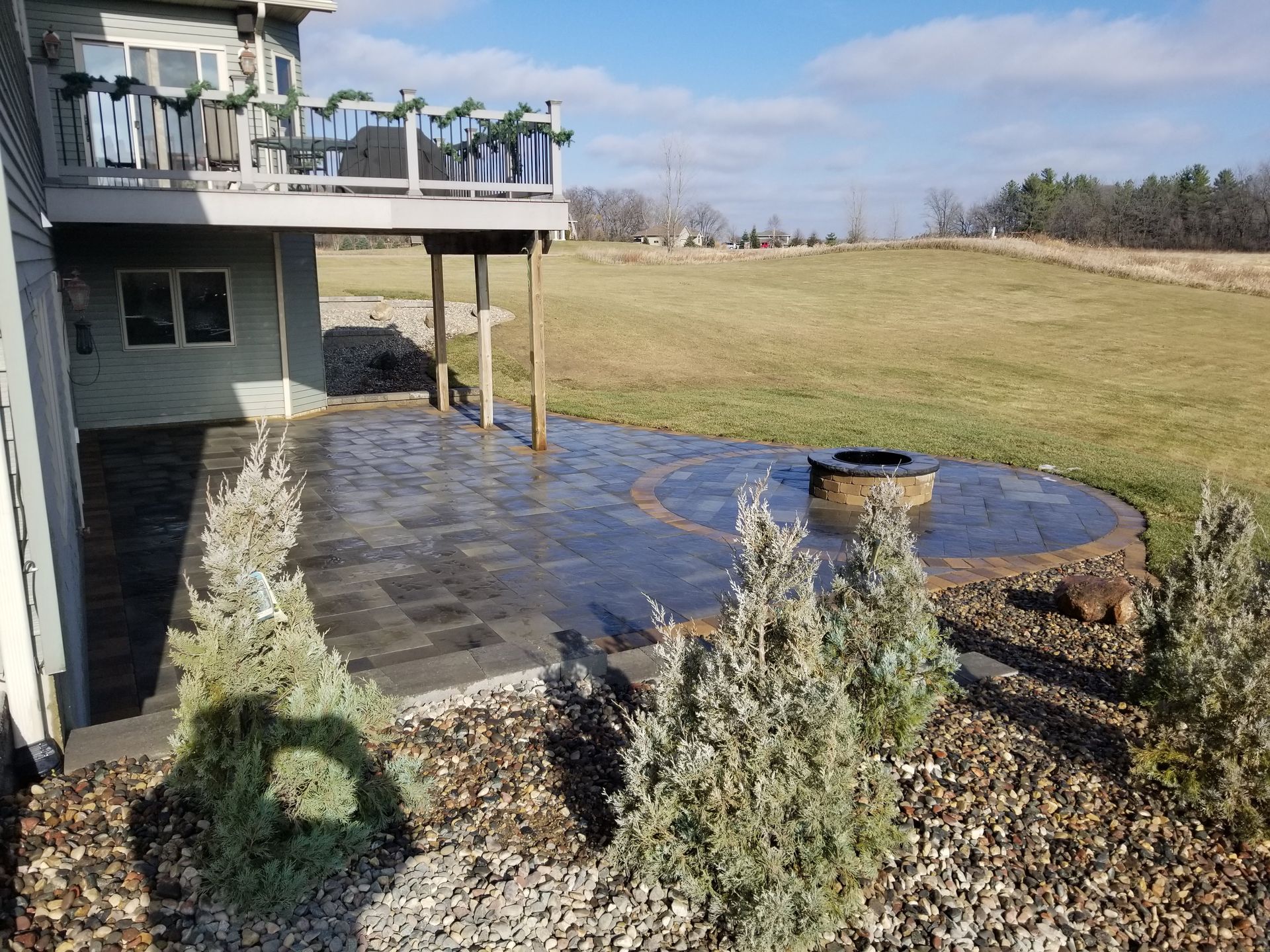A stone patio with a built-in fire pit sits beneath a wooden deck outside a house overlooking a grassy rural landscape.