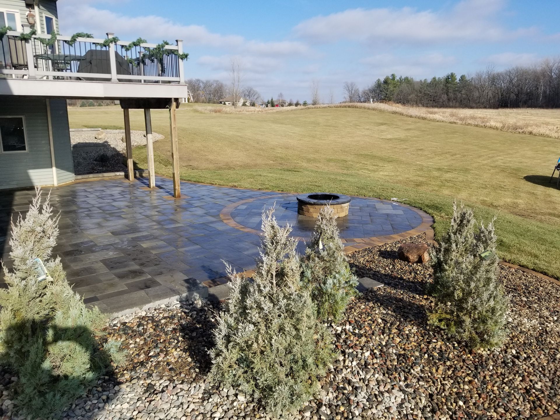 A stone patio with a fire pit sits under a deck overlooking a grassy, rural landscape under a clear, blue sky.